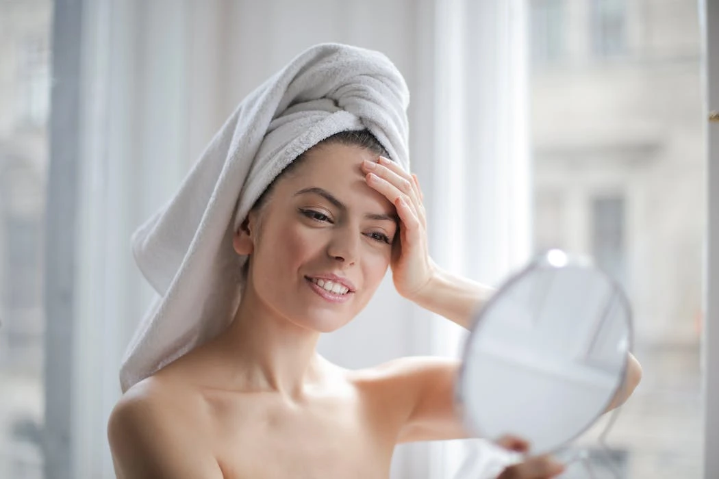 A woman with a towel on her head looking in a handheld mirror and smiling.